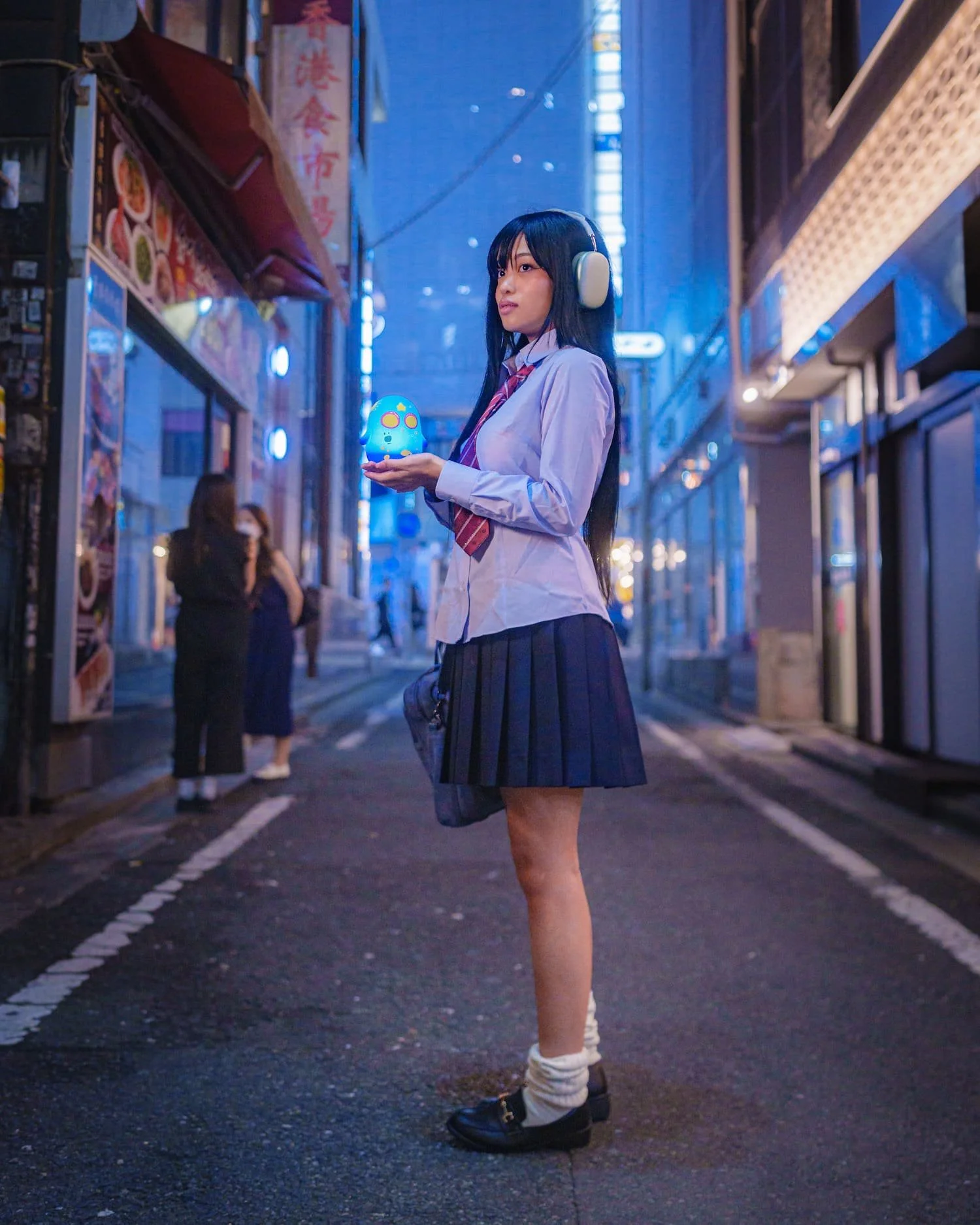 A young woman wearing a school uniform with headphones and black shoes is standing on a city street at dusk, holding a glowing blue cute character lamp.
