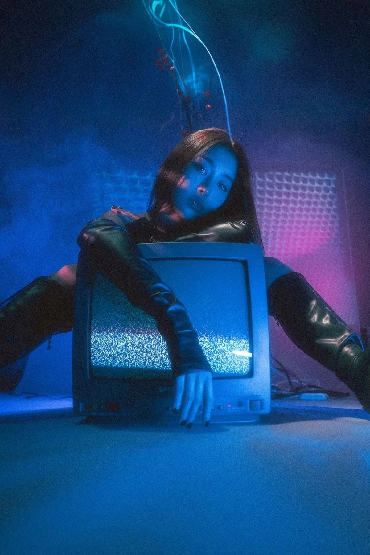 A woman with black hair in a dark room with neon blue and pink lighting, leaning on an old CRT monitor showing static on the screen, wearing a black leather outfit.