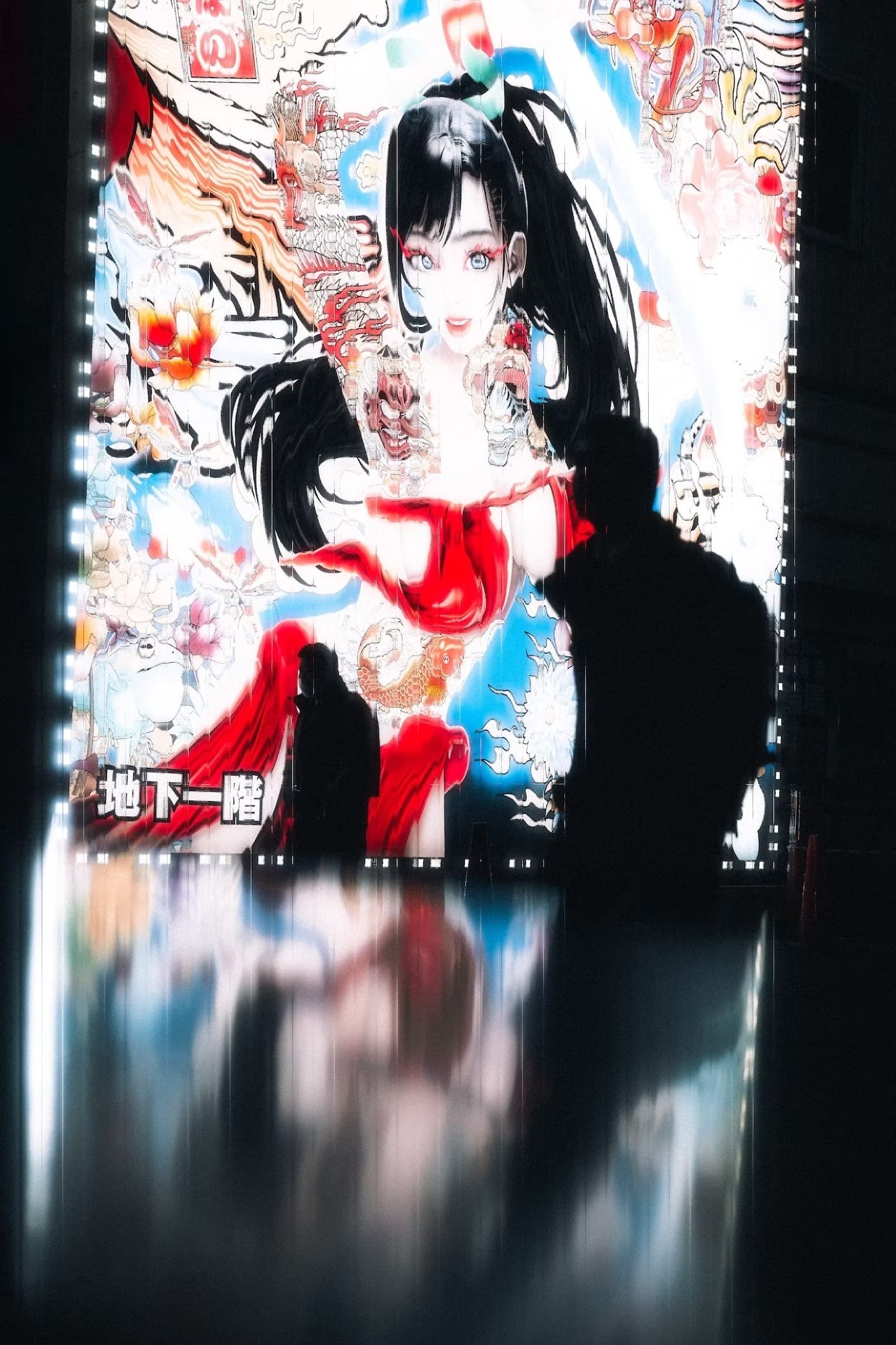 A large illuminated poster with artistic illustrations of a woman with long black hair, dressed in red, surrounded by colorful images and Japanese characters. The poster is reflected on a shiny surface below and has silhouettes of people looking at i