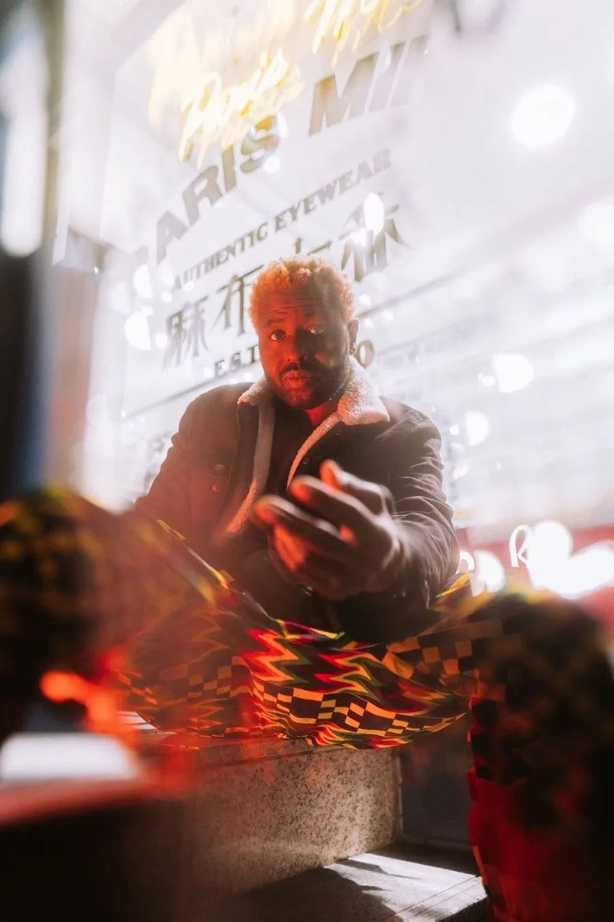 A man with light brown hair and a beard, wearing a dark jacket with a light-colored collar, looking into the camera while holding a mobile phone inside a brightly lit shop reflected in a mirror.