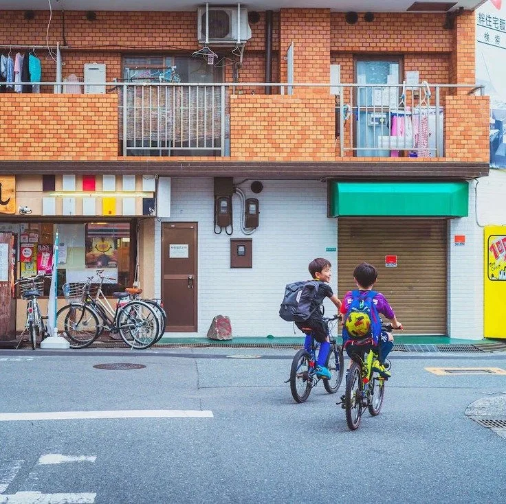 Two kids riding bicycles in front of a building in Osaka, captured while doing slow travel through the city.