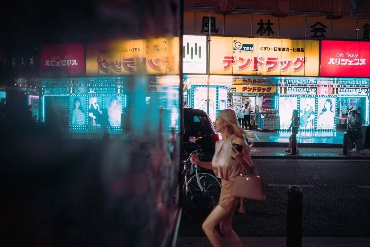 A woman with blonde hair in a beige trench coat and matching shorts, holding a purse and a smartphone, walking past a colorful, brightly lit storefront in an urban area at night, with neon signs and advertisements in Japanese. Kabukicho hostess snapp