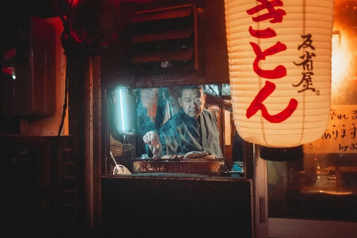 A man working at a food stand illuminated by a paper lantern with Japanese characters, in a dimly lit setting. Shot in Shimbashi by Tokyo street photographer Deniz Demir.