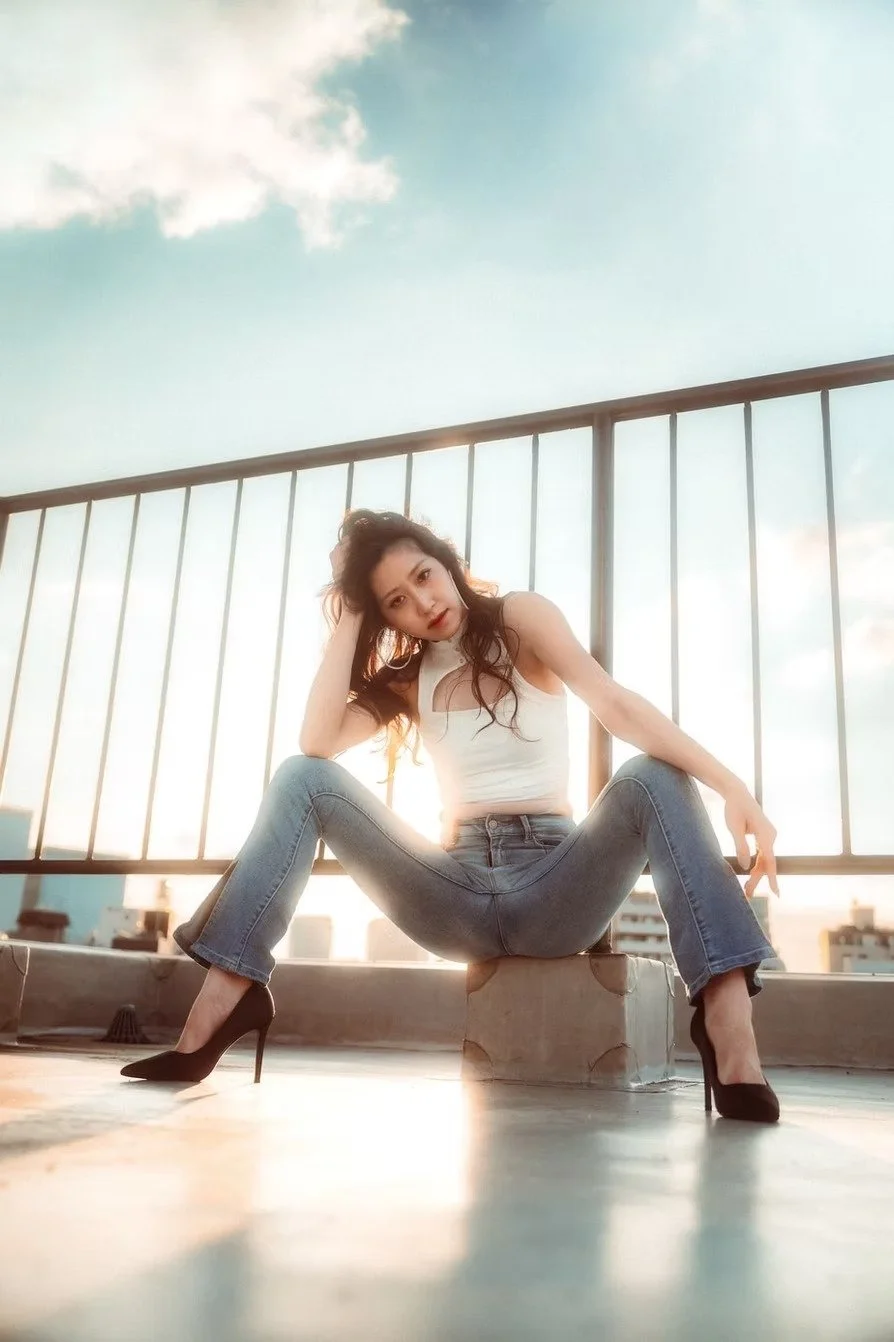 A woman with long dark hair in a white sleeveless top and blue jeans, sitting on a concrete block on a rooftop with a metal railing, during sunset with Tokyo cityscape in the background.