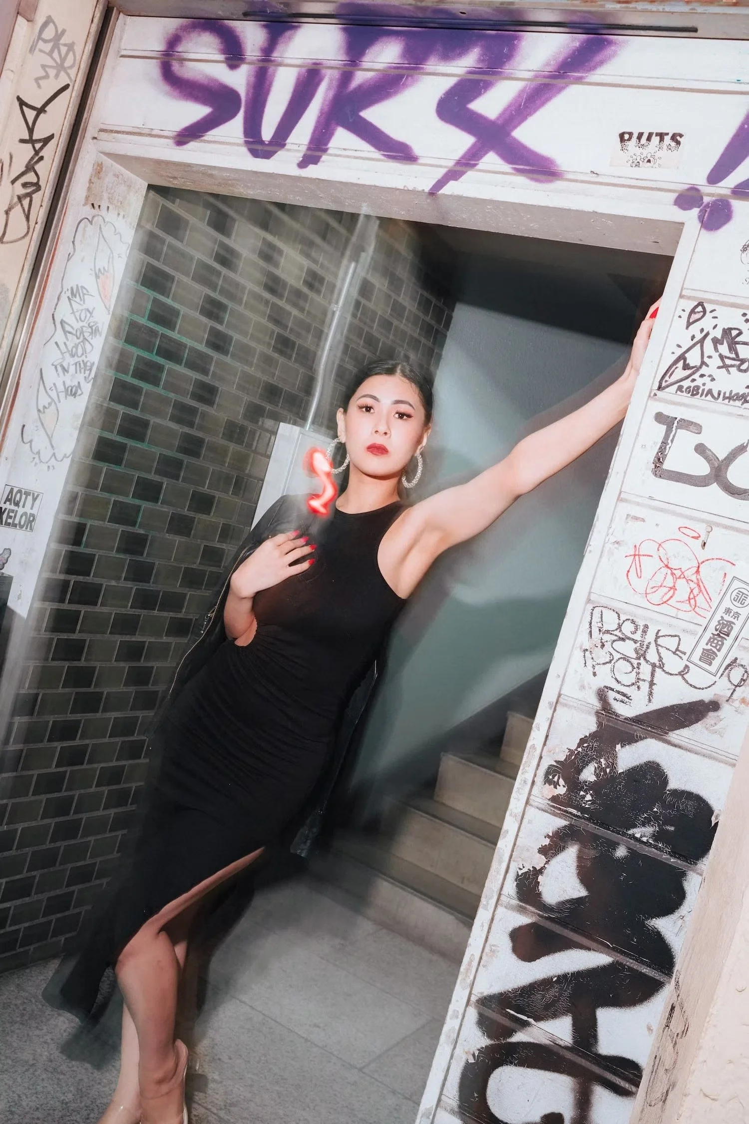 A woman in a black dress and red lipstick taking a mirror selfie in an urban setting. The mirror has graffiti and stickers, and she is holding her arm out with her hand near her chest. She has earrings and long, brightly colored nails.