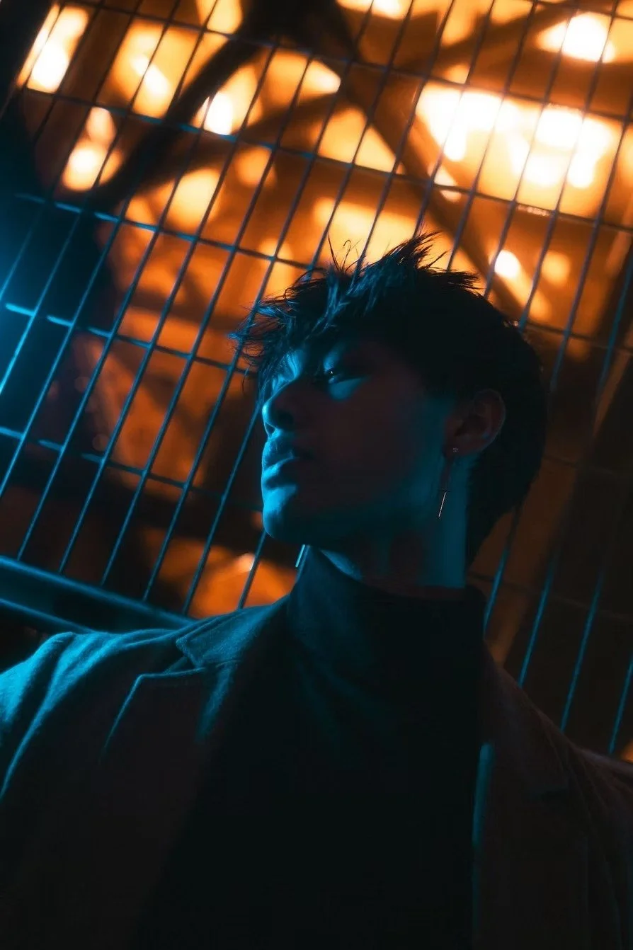 A person with short, spiky hair and an earring, looking to the side, illuminated by blue light, with a metal fence and orange, blurred lights in the background. Captured by Tokyo portrait photographer Deniz Demir.
