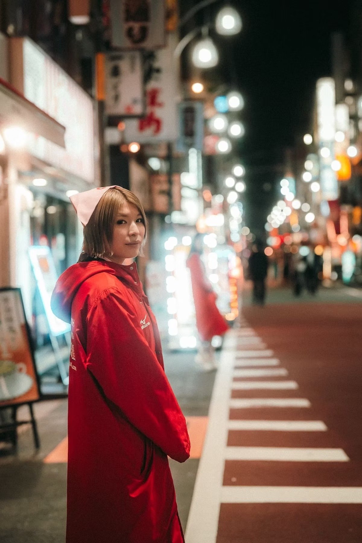 A woman in a red jacket with a pink cloth on her head standing on a city sidewalk at night with blurred streetlights and signs in the background shot in Shimbashi Tokyo.
