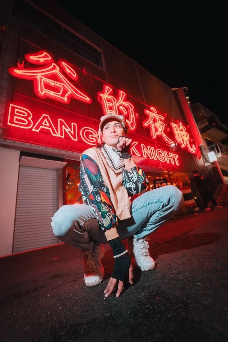 Person crouching in front of a neon sign that reads 'Bangkok Night' in Kanji and English