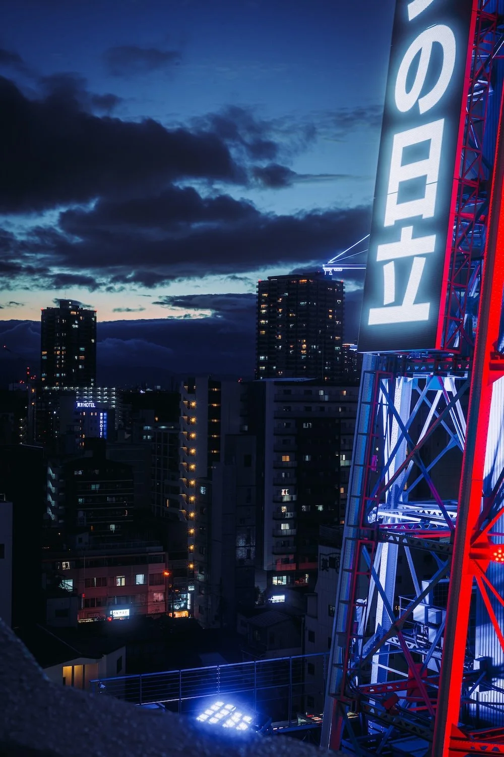 An Osaka city skyline at dusk with illuminated buildings and a large red and white neon sign reading 'LATTEA' on a tower structure shot in Shinsekai district by Deniz Demir.