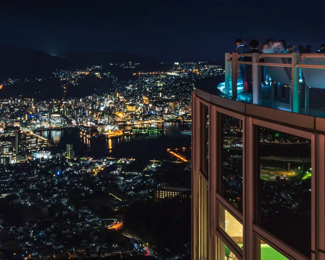Night view of a city skyline with illuminated buildings and a river, taken from a high observation deck with glass railings and several people sightseeing.