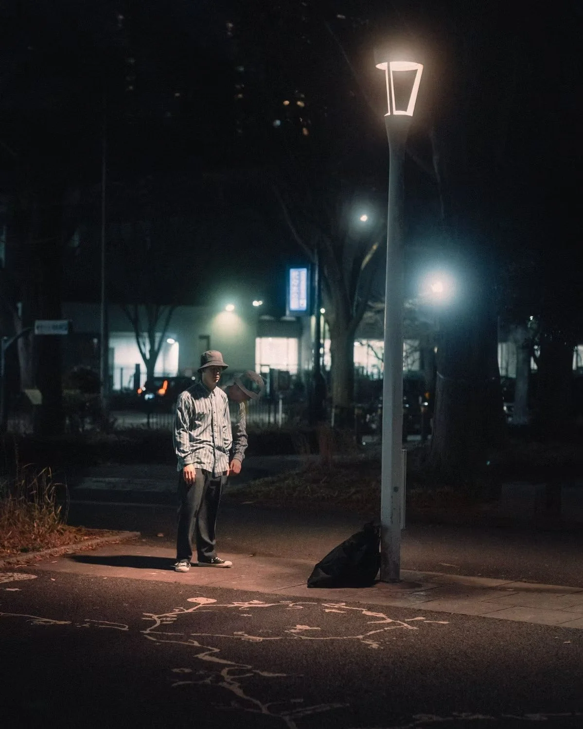 A person wearing a hoodie and hat standing on a dimly lit street at night near a streetlamp, with another person beside them. The background includes trees, buildings, and streetlights, with faintly visible parked cars.