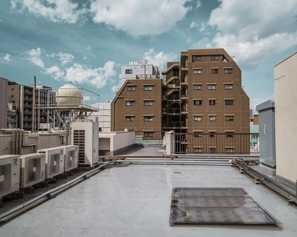 View from a building rooftop showing air conditioning units, vents, and a water tank with a city skyline of brown brick and gray buildings under a partly cloudy sky.