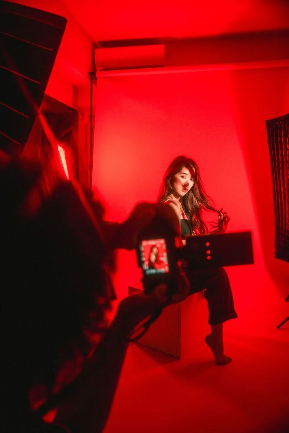 A model poses confidently in a dark environment with red and blue lighting illuminating her form. The lighting accentuates her sensual pose and the contrasting textures of her outfit, creating an intense and alluring visual.