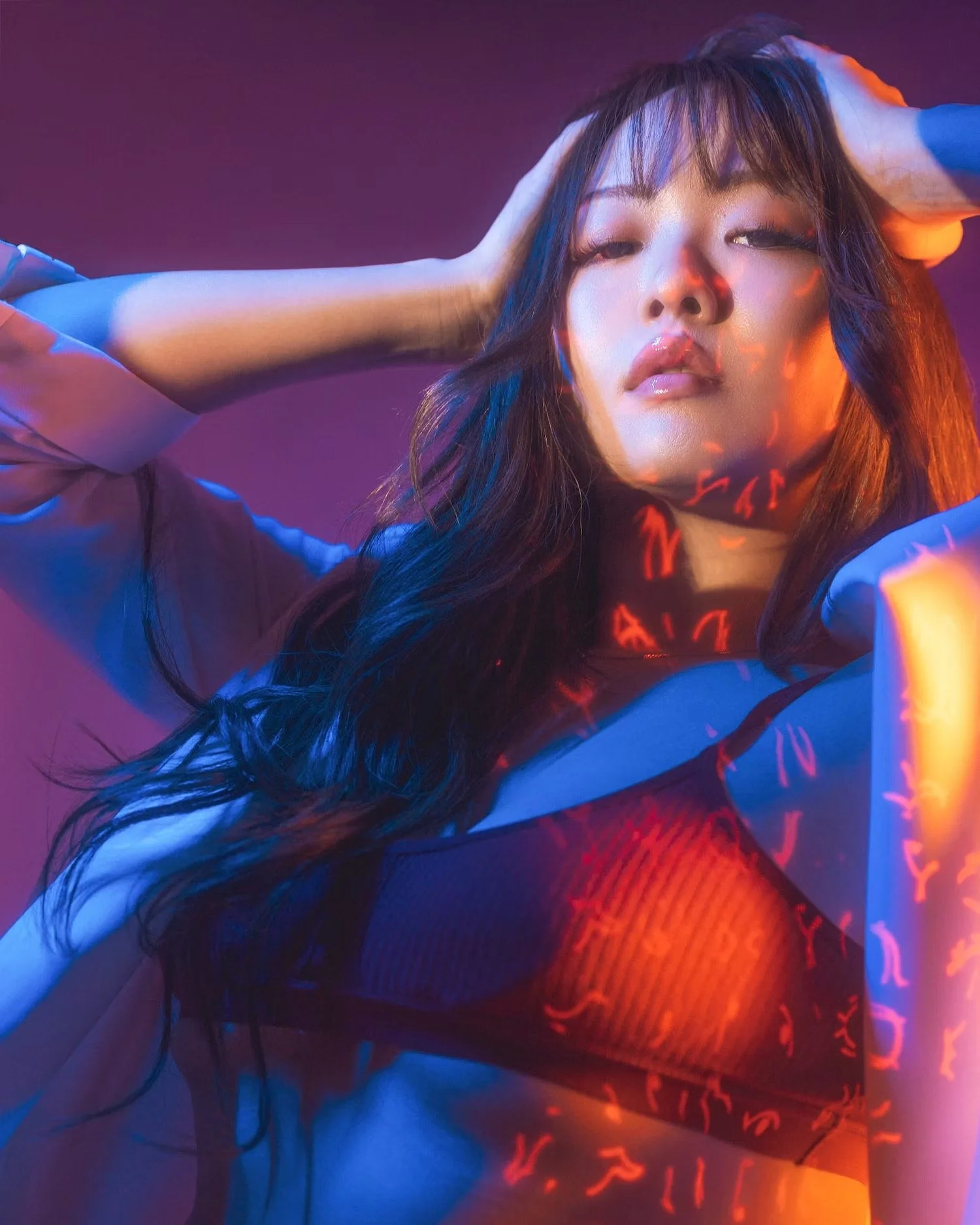 A woman with long dark hair posing with hands behind head, wearing a light blue shirt and black fishnet stockings, illuminated by colorful lights creating a vibrant, artistic atmosphere.