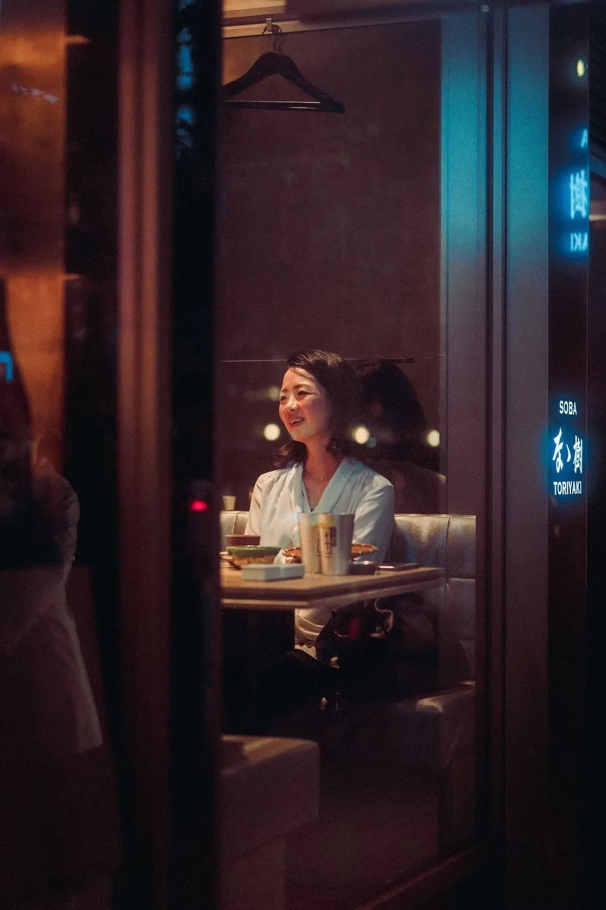 A woman with dark hair smiling, sitting at a dining table in a dimly lit restaurant, with plates and cups on the table, viewed through a partially open door or window. 
