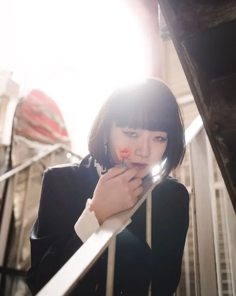 A woman with black hair and bangs applying makeup in front of a mirror with sunlight shining behind her.