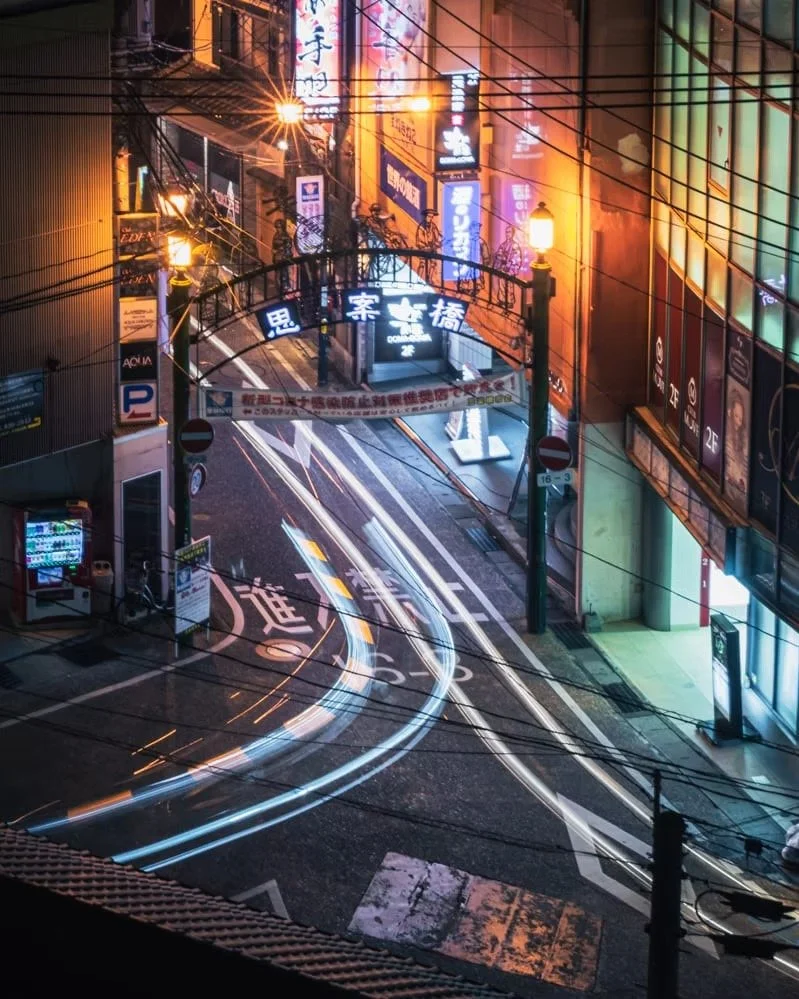 Nighttime city street scene with neon signs, streetlights, and vehicle light trails from moving cars.
