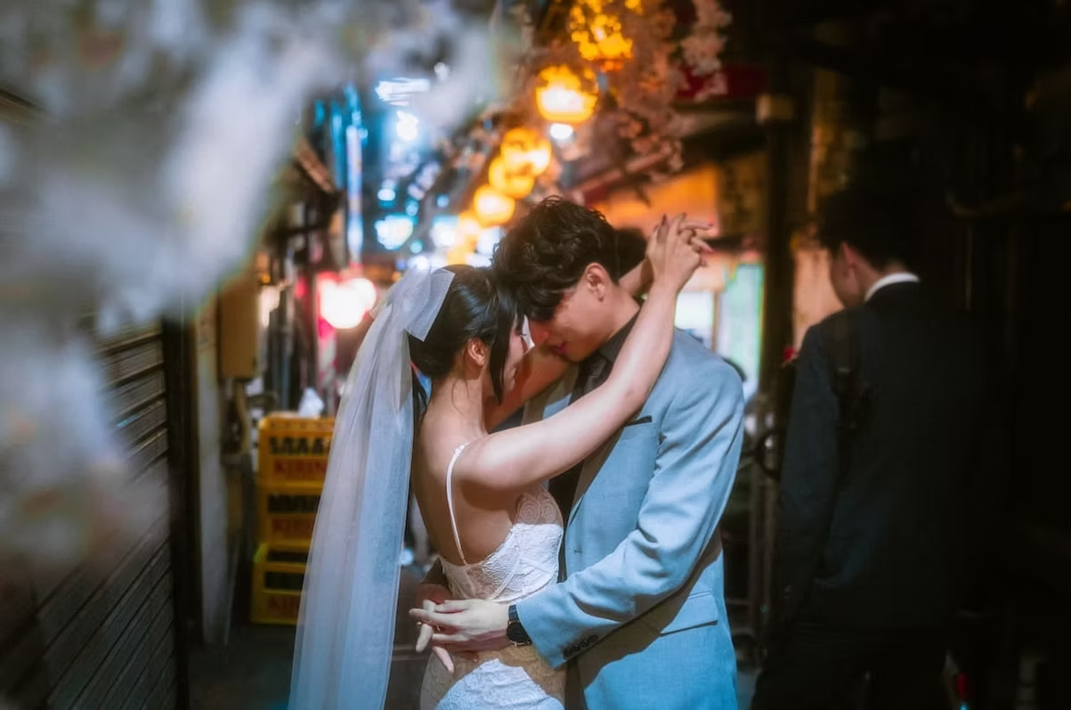 A bride and groom embrace in a narrow, lantern-lit alley with blurred background of hang lanterns and signs, suggesting a romantic moment.