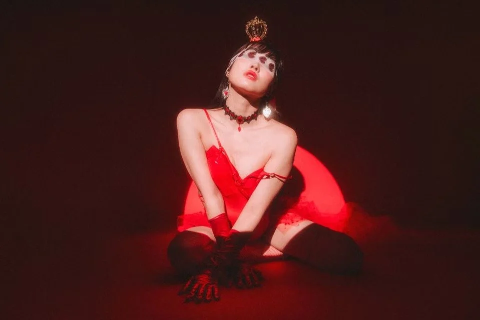 A woman dressed in red lingerie and gloves sits gracefully in a powerful, seductive pose. The bold red backdrop and dramatic lighting enhance the striking and confident atmosphere.