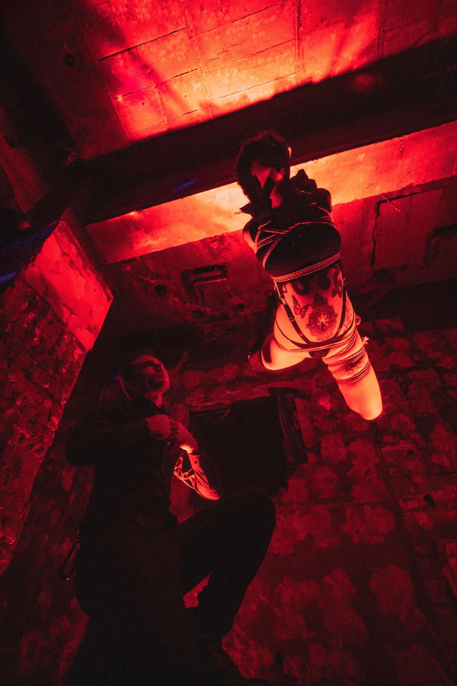 Bondage Ball 
---
A person with curly hair, wearing a painted shirt, is hanging upside down in a dimly lit room with red lighting. Two people are observing, one of whom is holding a phone, in a room with stone walls and a low ceiling.