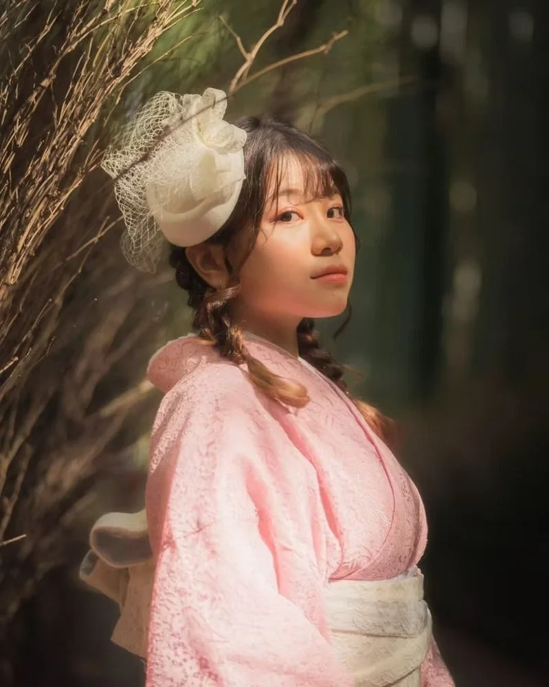 A young woman in a pink kimono with a white obi, standing outdoors near a cluster of brown reeds or dried branches, with soft lighting and a blurred green background.