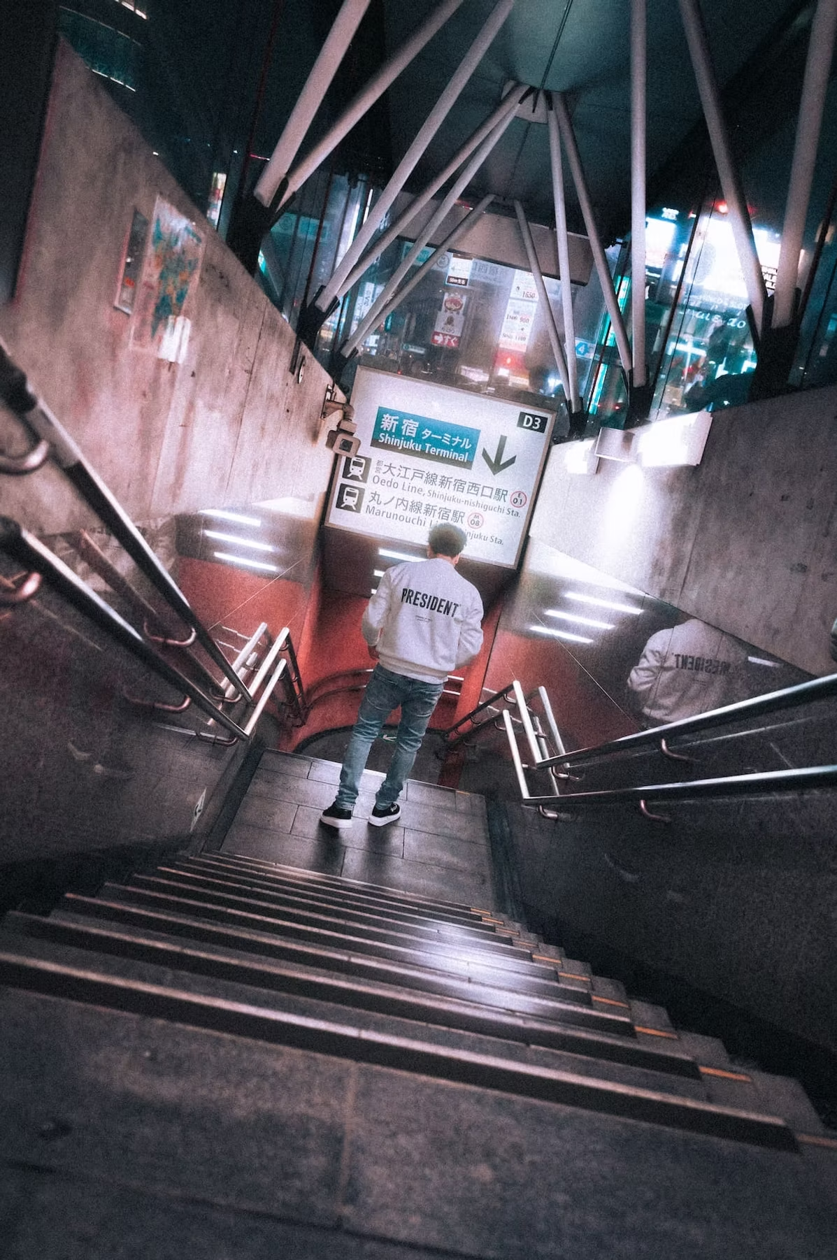 A person in a white shirt and jeans stands on the stairs leading down to an underground area, surrounded by a modern architectural design. The scene is lit by soft lighting.