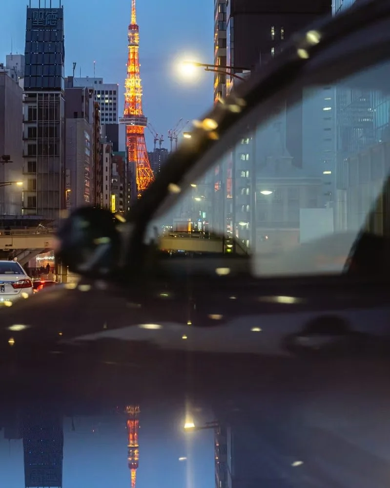 A nighttime cityscape featuring Tokyo Tower illuminated in orange and yellow, surrounded by tall buildings with construction cranes in the background, seen through the windshield of a vehicle with reflections on its surface.
