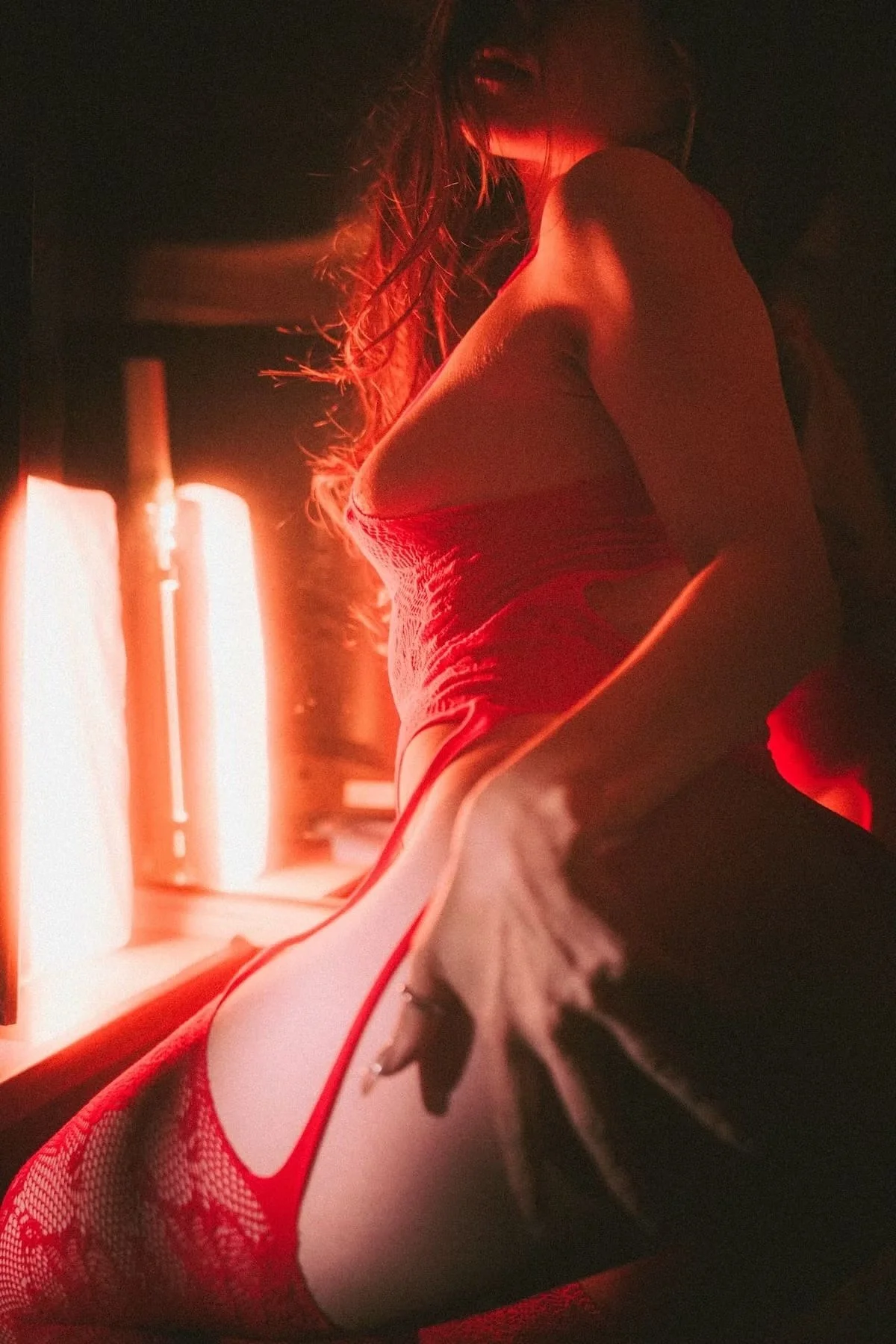 A woman dressed in red lingerie and fishnet stockings sitting in front of red incandescent lights, smiling with her head tilted back and hair flowing.