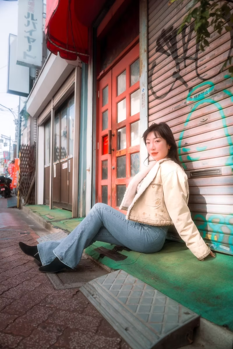 A young woman with dark hair sitting on a sidewalk against a graffiti-covered metal shutter in an urban area, wearing a beige jacket, light blue jeans, and black shoes.