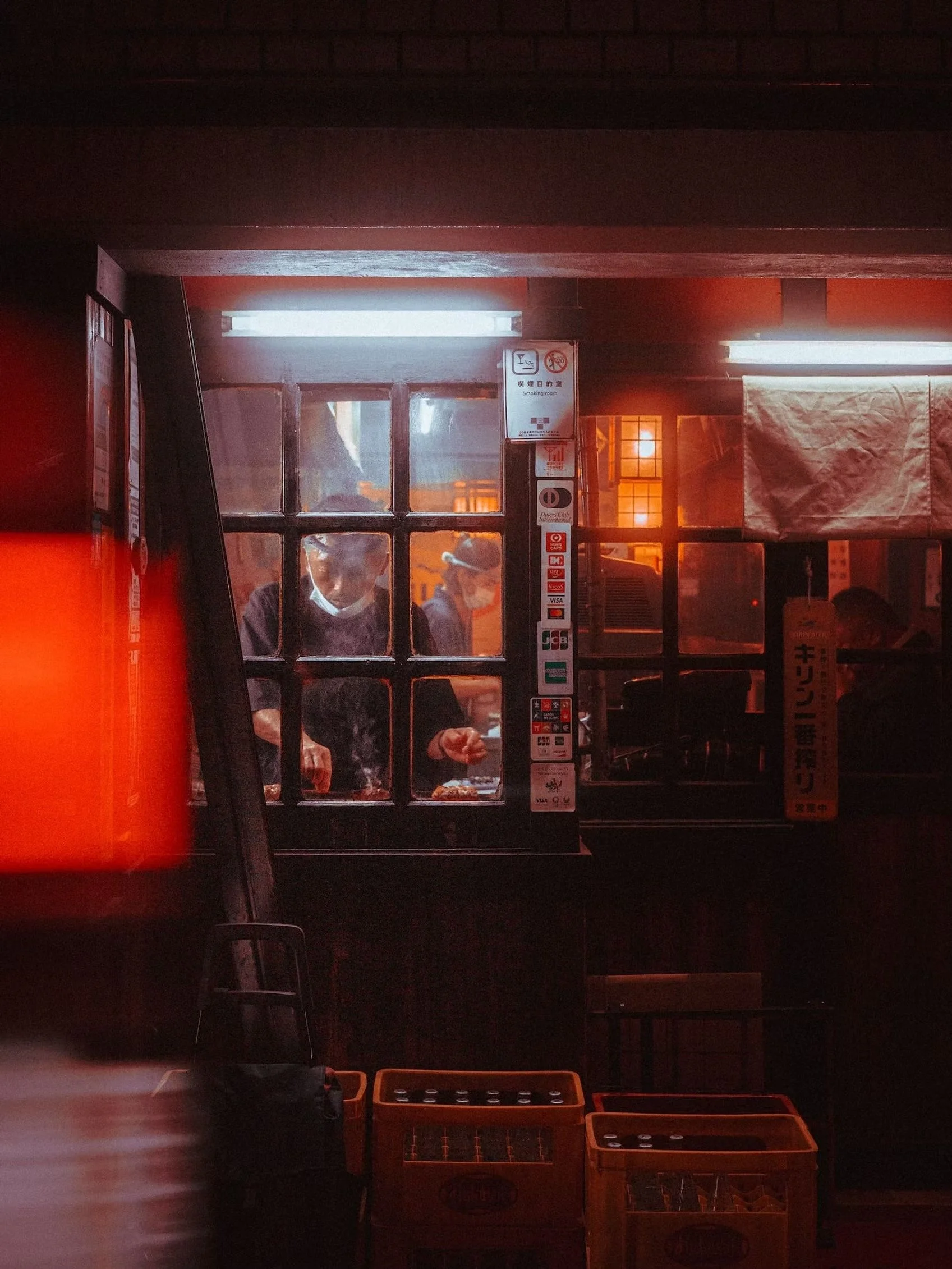 A dimly lit indoor scene with a window, a man wearing glasses and a mask, various signs, and a few orange baskets on the floor.