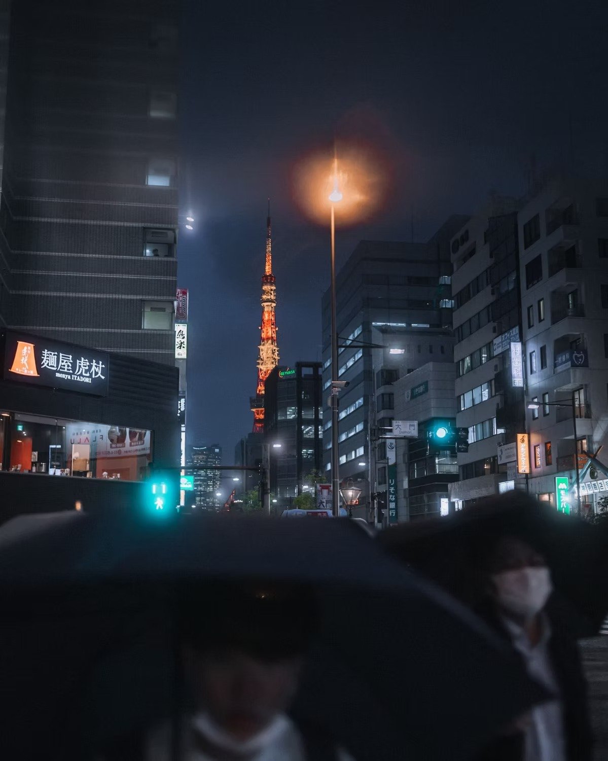 Nighttime city street scene in Tokyo with illuminated buildings and the Tokyo Tower in the background. People wearing masks are visible in the foreground.