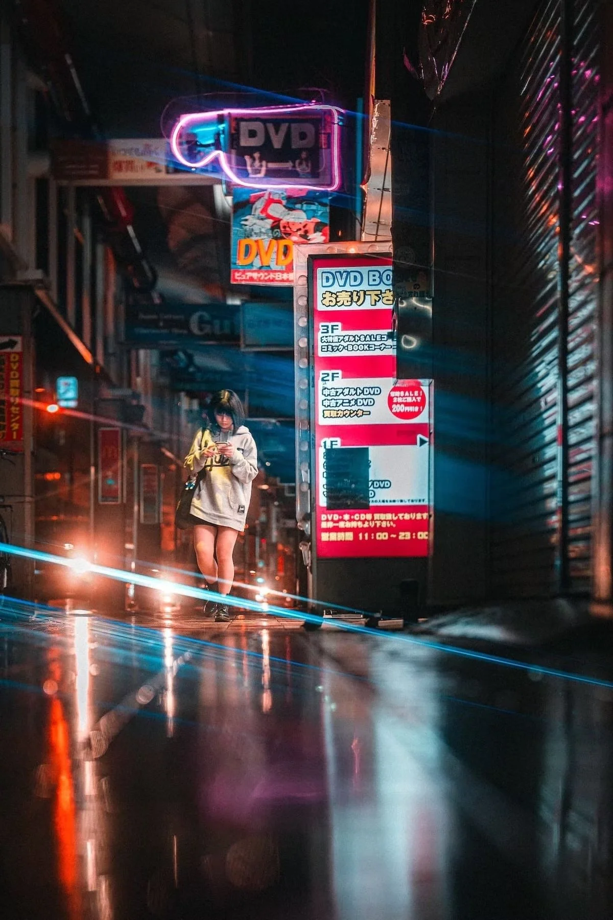 A young woman in a hoodie and shorts walking on a wet city street at night, illuminated by neon signs and streetlights, with reflections on the pavement. Osaka night scenes shot by Tokyo street photographer Deniz Demir.