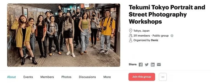 A group of diverse people pose in a Tokyo street under warm lighting, some holding cameras. The setting has an urban, artistic vibe, suggesting a workshop event.