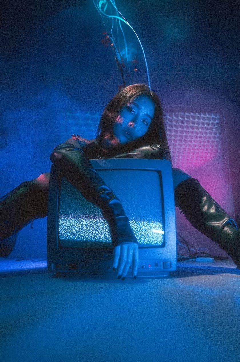 A woman in a sleek black outfit poses dynamically beside an old CRT television in a dark room. The neon hues and striking composition create a retro-futuristic cyberpunk look.