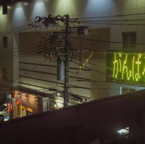 A glowing Japanese sign reading "がんばろう" (Let’s do our best) illuminates a night scene with tangled power lines and a dimly lit street.