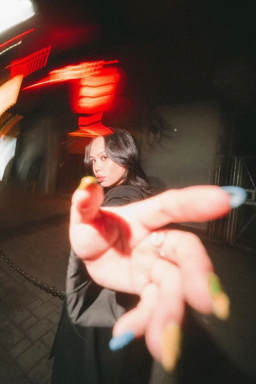 A woman with dark hair pointing towards the camera with her finger, in a dimly lit environment with red and orange light streaks in the background. Shot by Tokyo portrait photographer Deniz Demir.