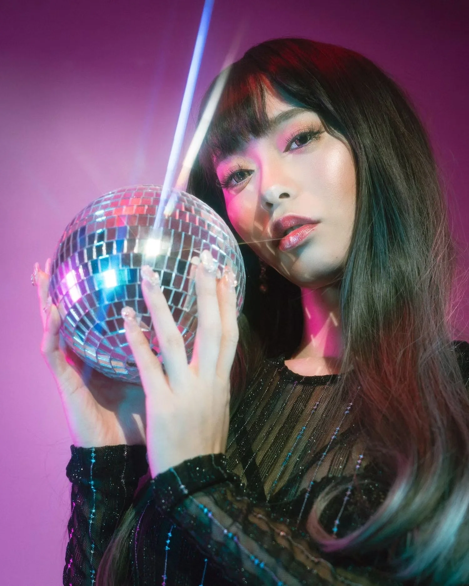 A woman with long dark hair and makeup holding a small disco ball with a bright light shining on it, against a purple-pink background.