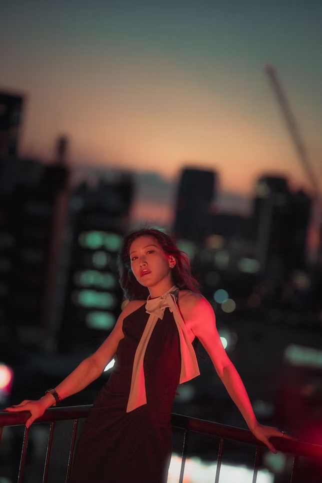 A woman standing on a balcony during sunset, city skyline in the background. She is wearing a black dress with a white scarf tied around her neck.