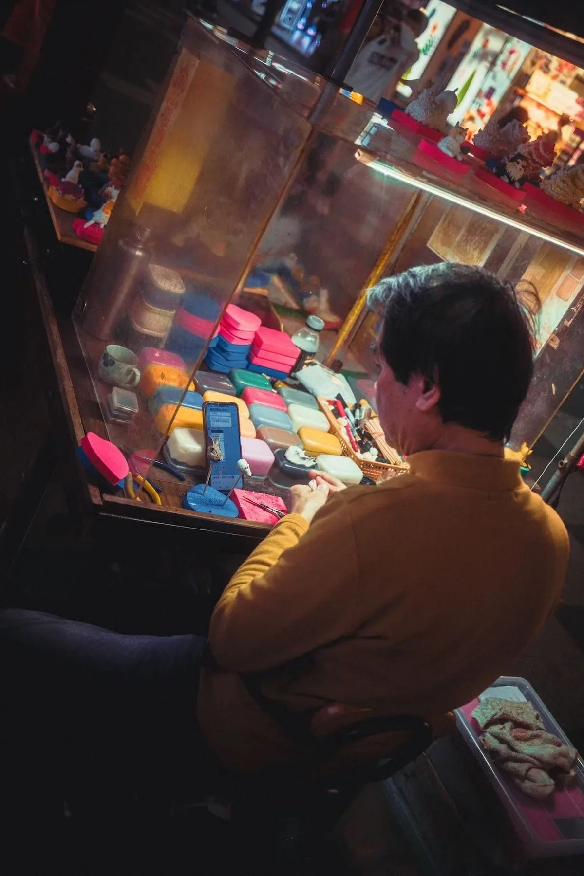 A person with dark hair and wearing a yellow shirt sitting at a booth in a market or shop, looking at various colorful boxes and containers behind a glass display case. Taiwan night market scenes by Deniz Demir.