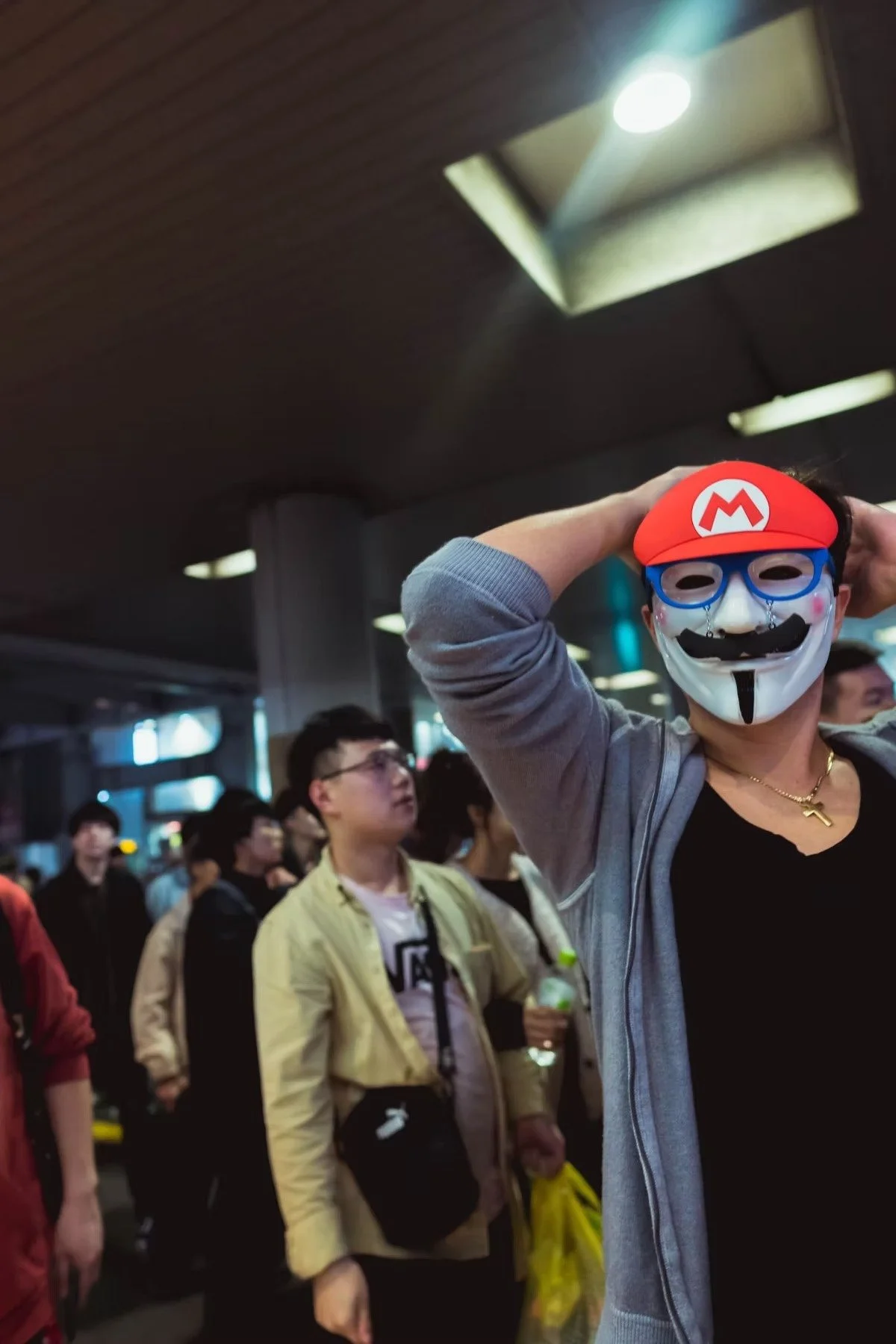 Person wearing a Guy Fawkes mask with a Mario hat and blue glasses poses with hands behind head in a crowded indoor space in Shibuya for Halloween shot by Tokyo street photographer Deniz Demir.