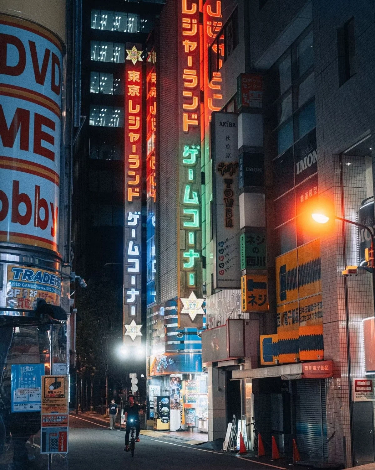 Night street scene in Tokyo with illuminated neon signs in Japanese, people walking and cycling, and a bright streetlamp on the right shot in Akihabara, Tokyo by street photographer Deniz Demir.