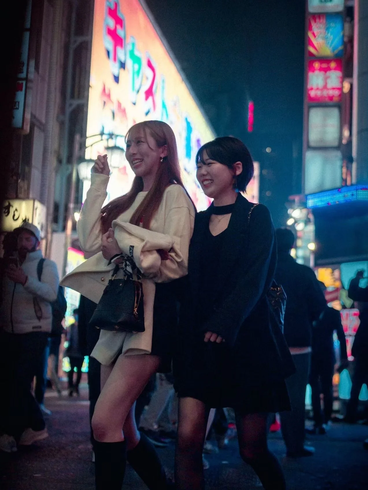 Two young women smiling and walking in a busy city street at night, illuminated by bright neon signs and displays.