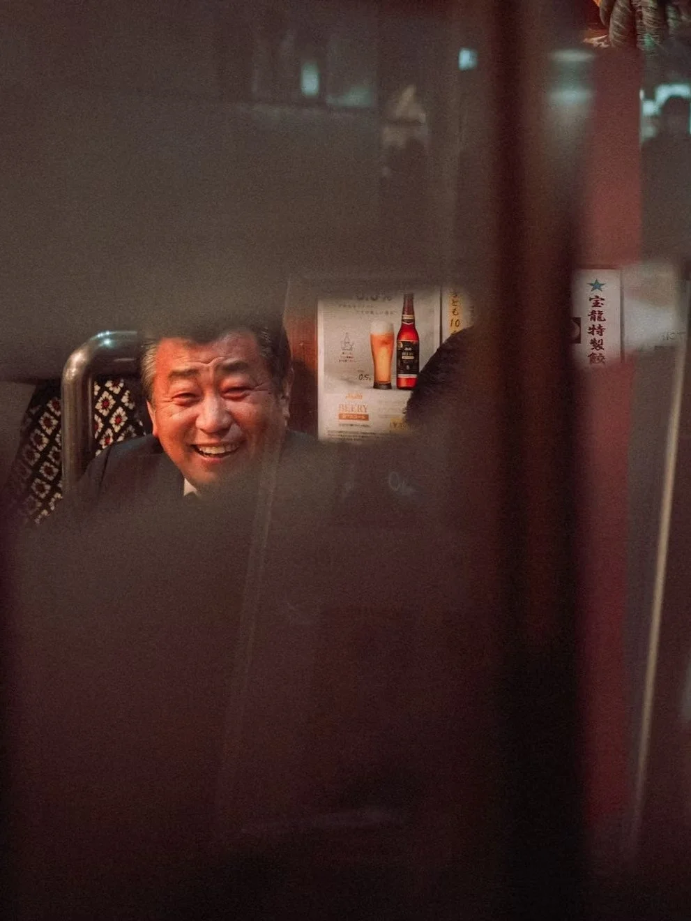 A person smiling and laughing, viewed through a gap with a dark foreground, in what appears to be a restaurant or bar setting.