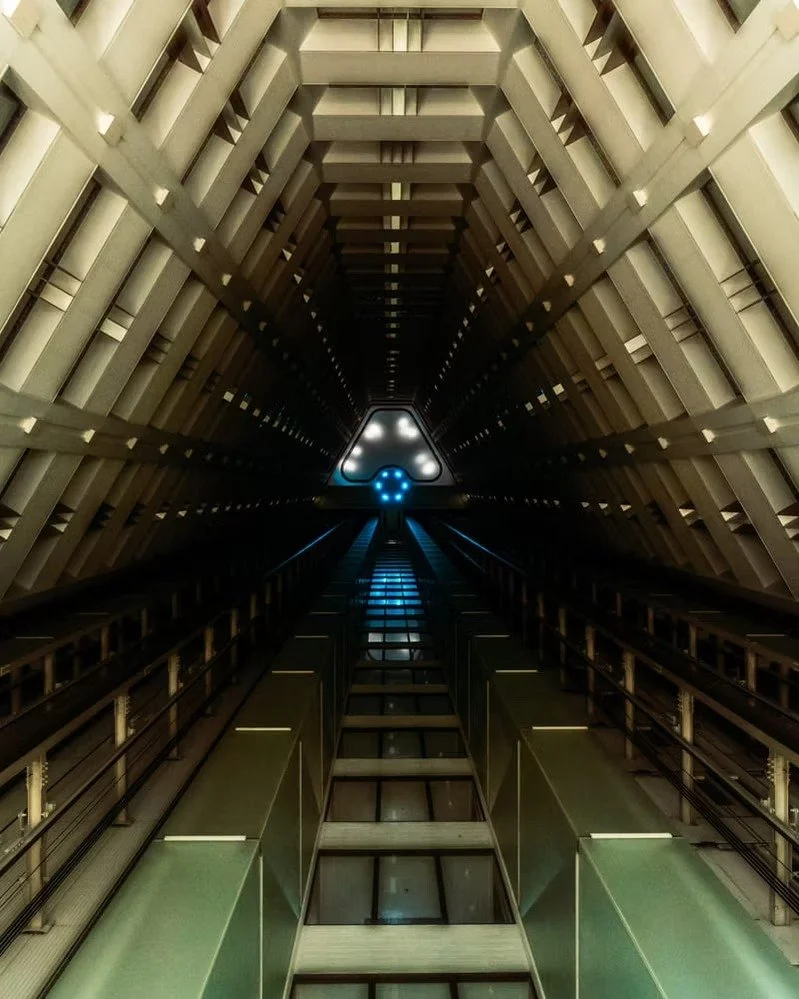 A view from the bottom of a modern elevator shaft looking up, showing the interior structure, railings, and a blue circular light at the top.