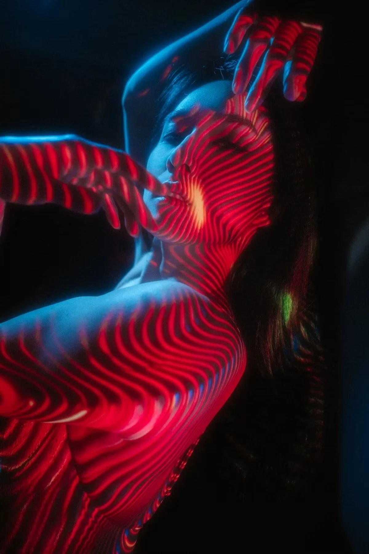 A woman in a black hood with red and blue neon light patterns projected onto her face and body, creating a colorful, abstract visual effect.