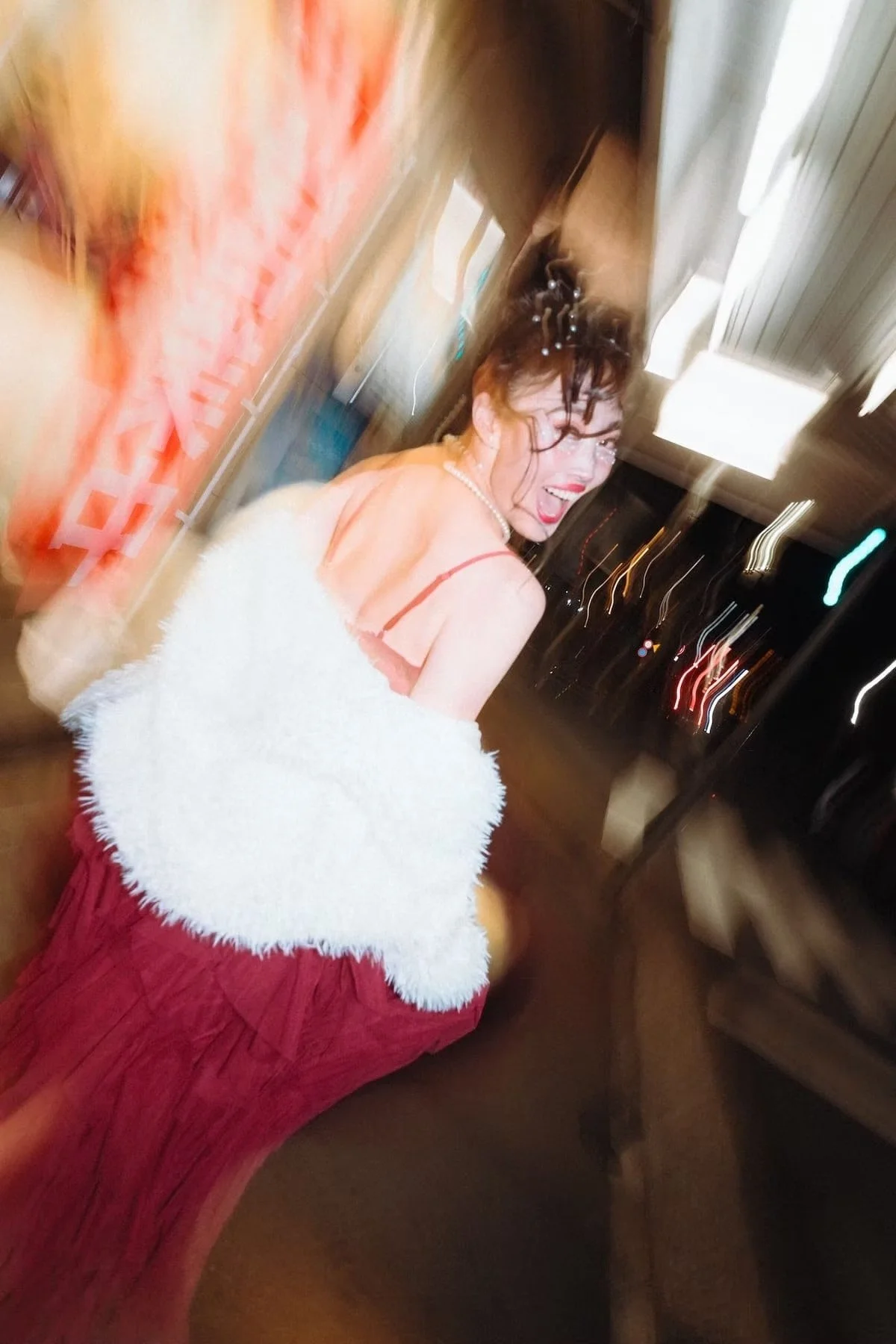 A beautiful woman in a pink dress with spaghetti straps, wearing a pearl necklace, is smiling and laughing in a dimly lit room. She has curly hair decorated with hairpins and is holding a white fluffy shawl. Deniz Demir portrait photographer.