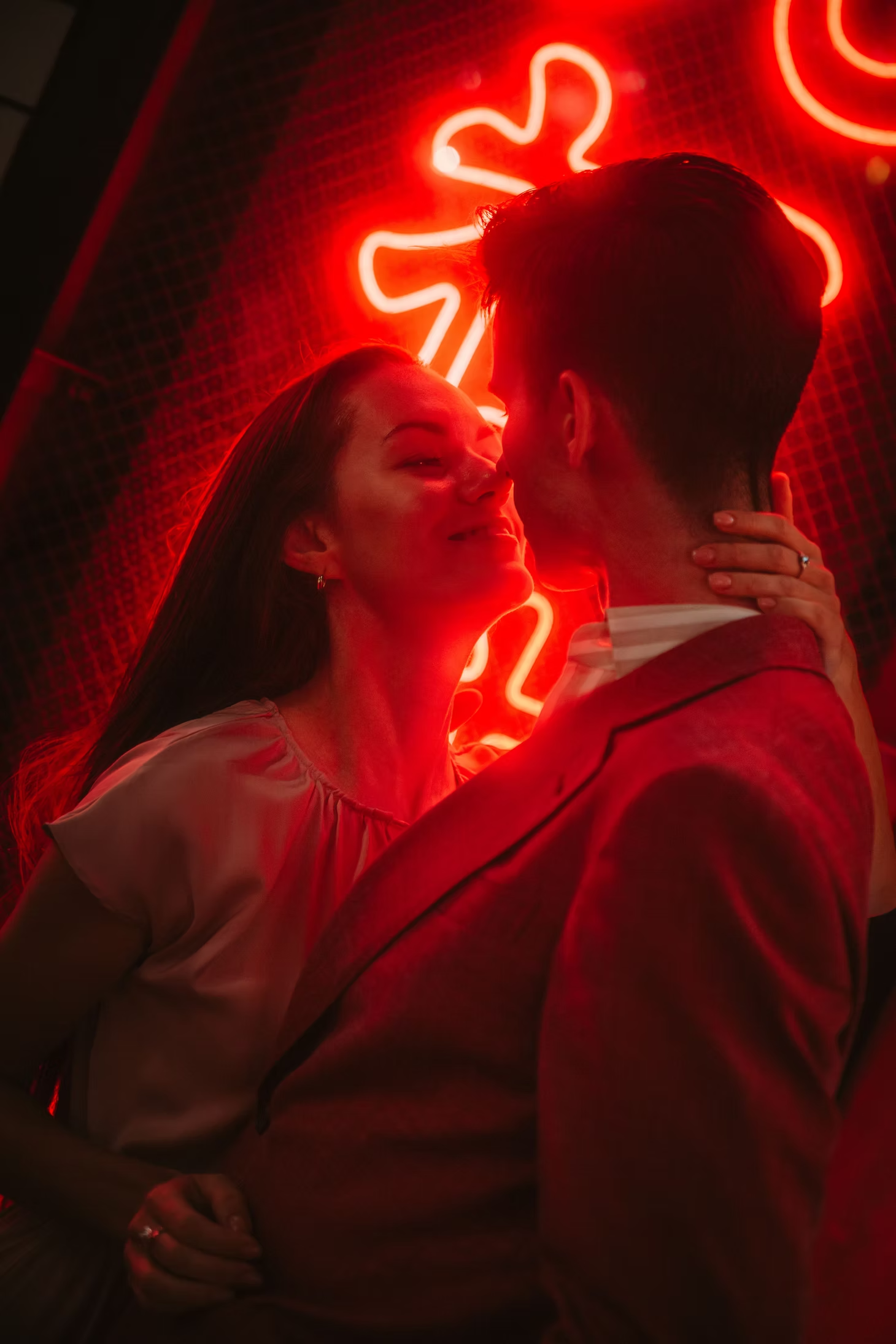 A woman and man sharing a close, intimate moment in front of a red neon-lit background featuring a gingerbread man and heart shapes.