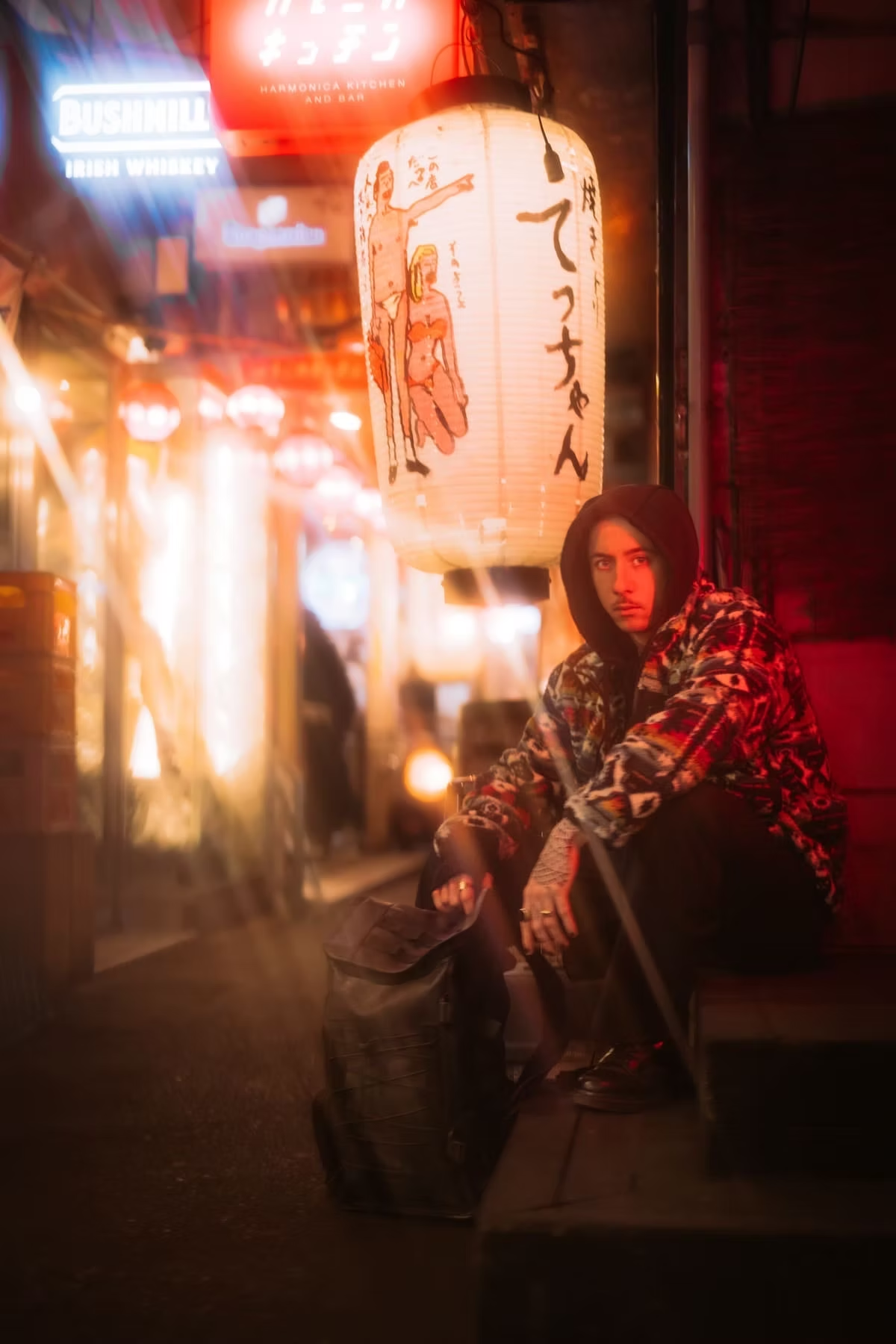 A man in a hoodie and patterned jacket sits against a wall with a backpack in a dimly lit alley, surrounded by glowing lanterns and neon sign