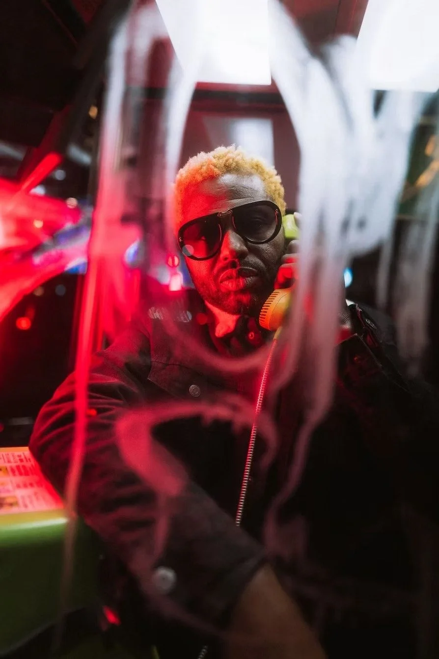 A man with blond hair and sunglasses, talking on a yellow retro phone, seen through a transparent plastic barrier, with red neon lights and blurred background.