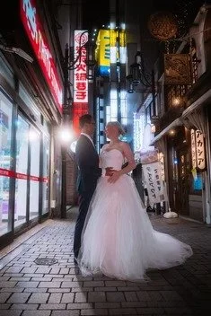 A couple dressed in wedding attire stands in a narrow Tokyo alley illuminated by bright neon signs. The groom in a suit and the bride in a wedding dress share an intimate moment, surrounded by the lively atmosphere of the city.